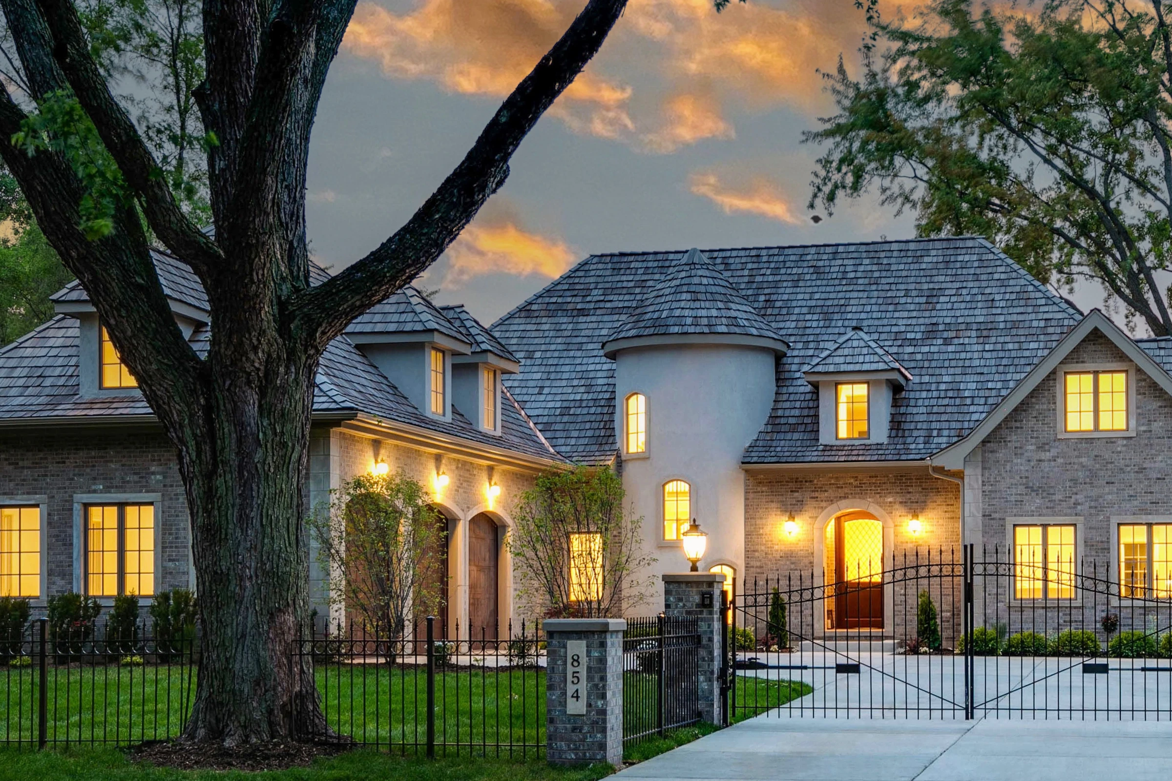 A stone residence by L'Atelier Elena glowing at dusk, gated approach lit beneath a sunset sky